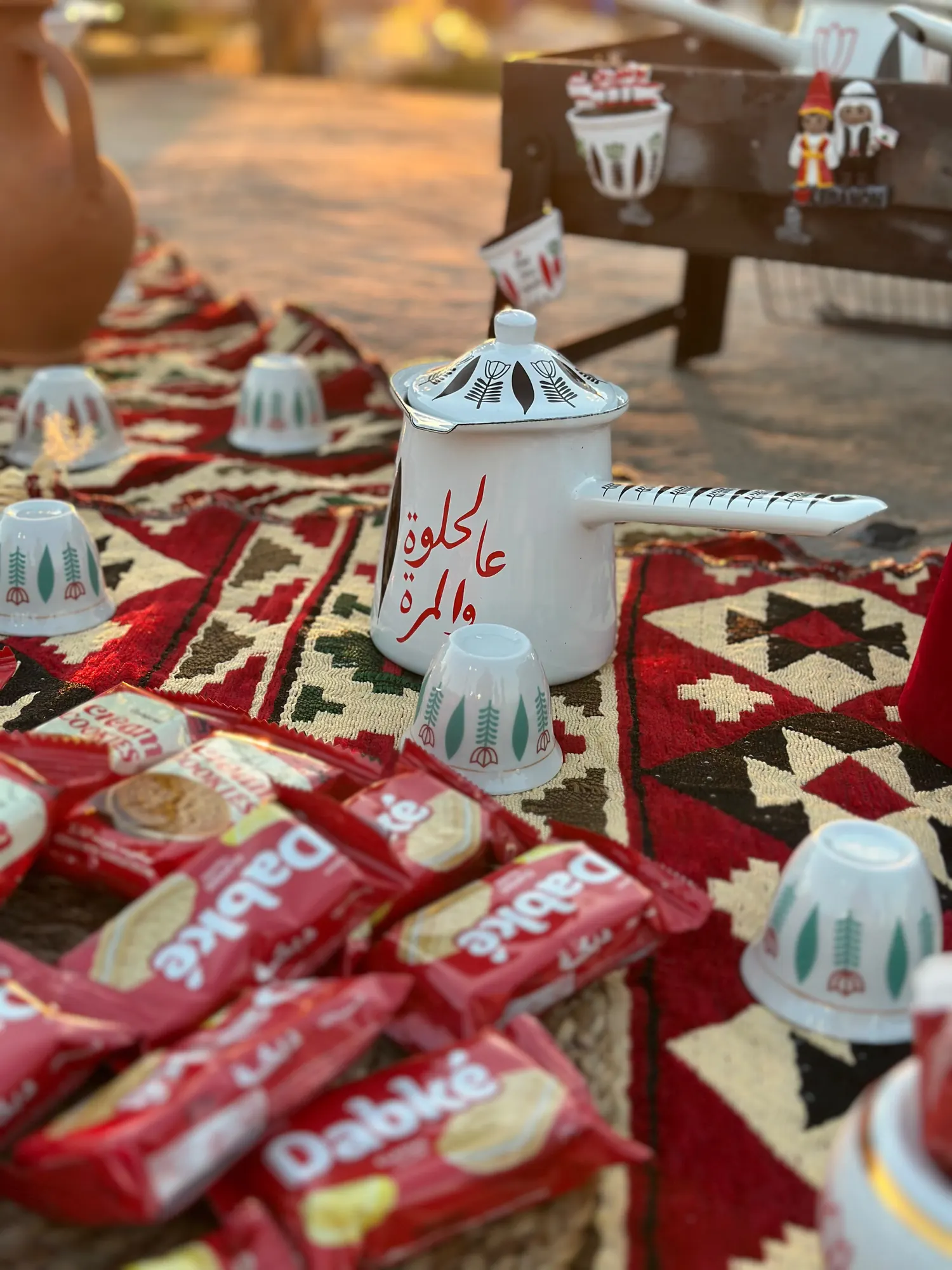 An Arabic coffee setup at dusk with the pot, cups and wrapped biscuits on a woven kilim